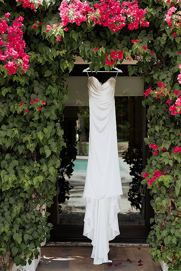 Wedding dress hanging on a hanger, a strapless gown with draped bodice and sweetheart neckline framed by a vine arch and doorway with pink bougainvillea