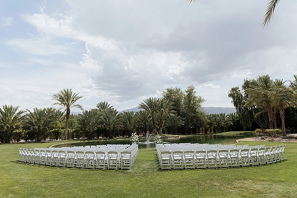 Ceremony setup for an outdoor wedding ceremony with curved white folding chairs and white floral arrangements by a pond and fountain with mountains beyond