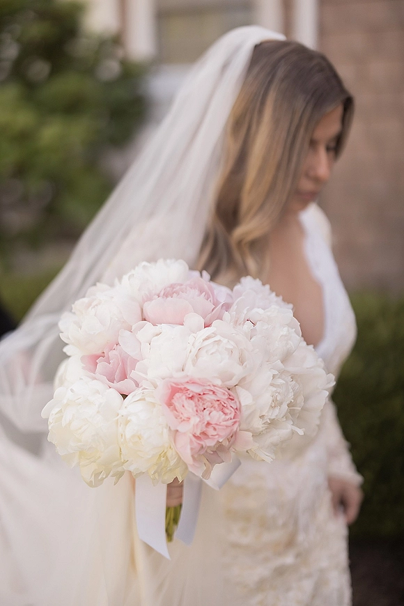 Bridal bouquet of white peony bouquet blooms in blush and ivory, tied with ribbon, held against lace dress and veil by stone wall
