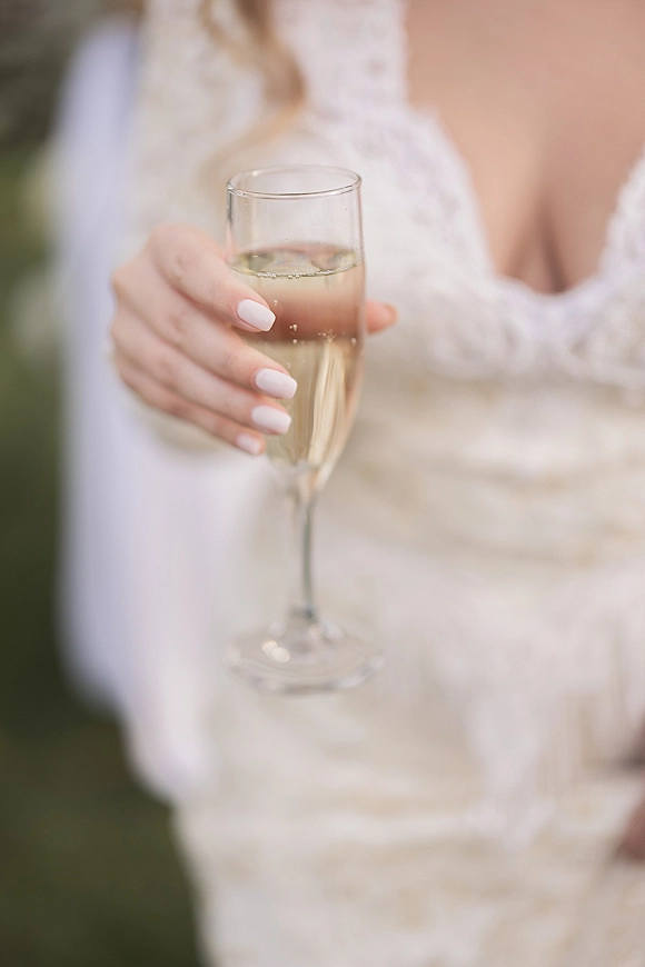 Champagne toast as bride holding champagne glass in a sparkling flute, lace sleeves and French manicure visible against a soft blurred backdrop