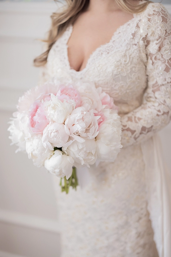 Bridal bouquet of pink and white peonies held against a lace long-sleeve V-neck wedding gown in front of a light neutral wall
