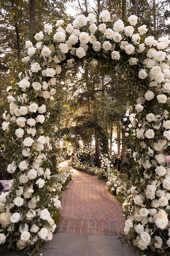 Ceremony aisle decor with a wedding floral arch of white roses and greenery garlands framing a sunlit brick walkway in a garden setting