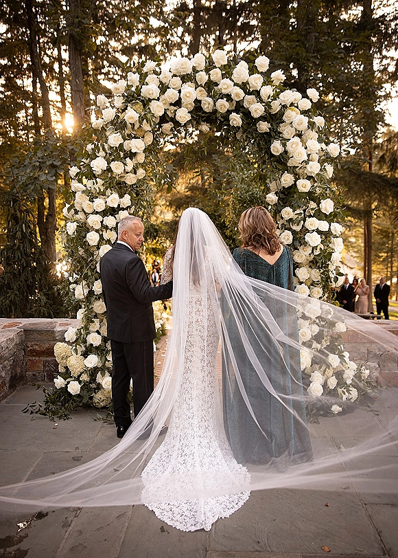 Wedding processional as bride walking down aisle from behind in lace dress and cathedral veil toward a white rose arch on a sunlit garden terrace