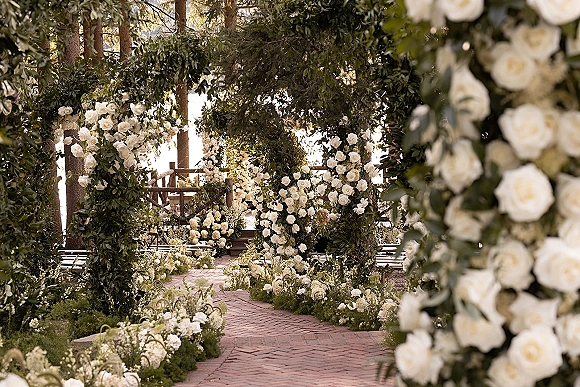 Ceremony aisle decor with a white rose wedding arch and greenery garlands lining a brick walkway, with wooden benches and sunlit trees
