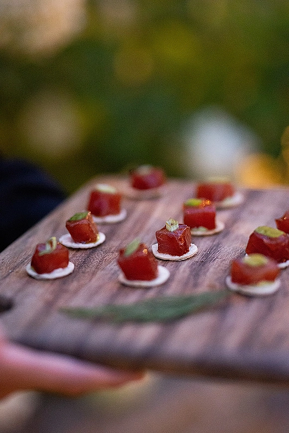 Wedding appetizers on a wood serving board, featuring bite-size tuna tartare canapes on crackers with green garnish and blurred garden lights