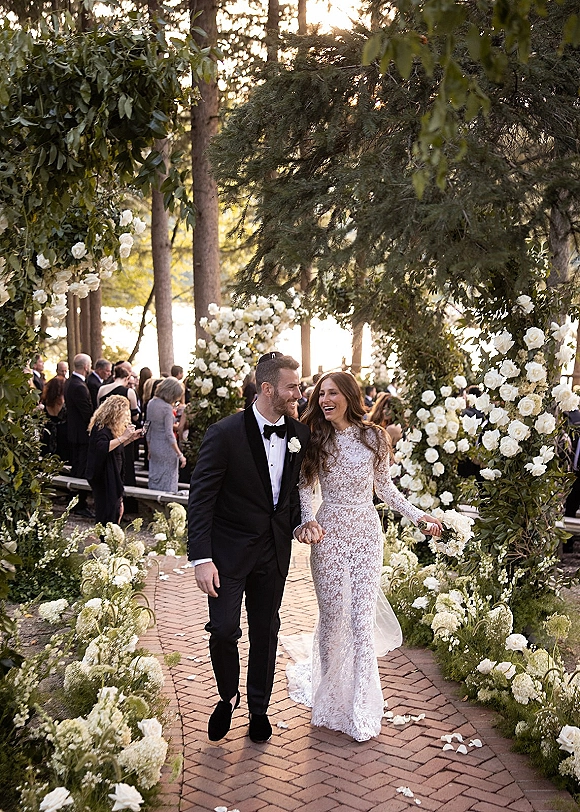 Wedding recessional as bride and groom walk the aisle laughing, her lace gown and bouquet framed by a white rose arch in sunlight