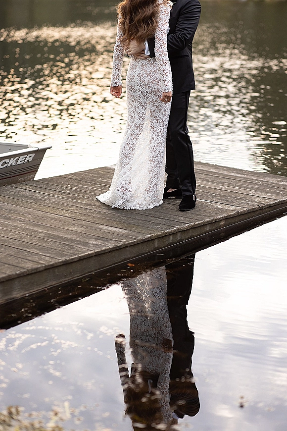 Couple portrait of bride and groom embrace from behind on a wooden dock by the lake, her long sleeve lace dress reflected in water