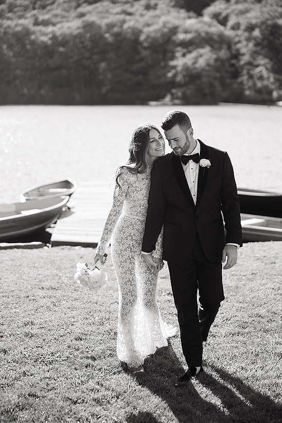 Couple portrait of bride holding bouquet in a long sleeve lace wedding dress with groom in tuxedo on a lakeside dock by rowboats