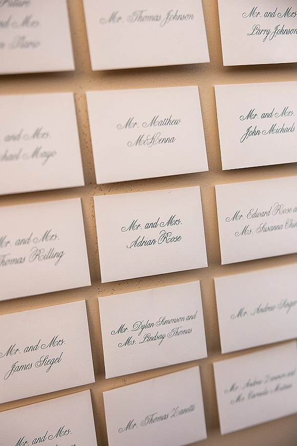 Wedding escort cards with calligraphy in black ink on white paper, neatly arranged in rows on a tan display board