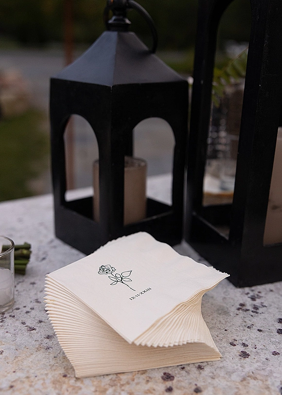 Wedding napkins and custom cocktail napkins with a rose illustration on a table beside black lanterns and candles along a greenery walkway