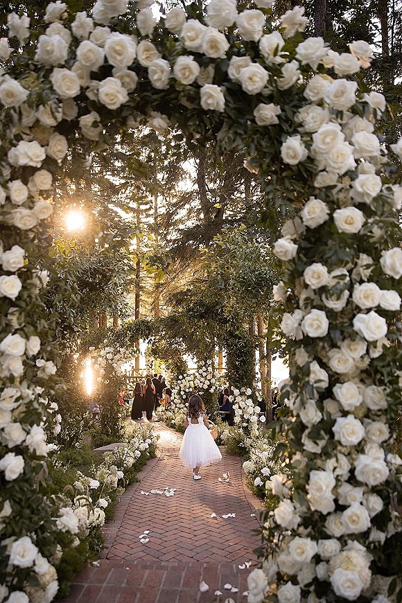 Ceremony aisle design with a white rose wedding arch, greenery garlands and aisle florals on a sunlit brick path beneath tall trees