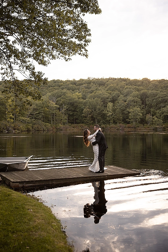 Couple portrait on a wooden dock, bride in a lace wedding dress dipping with groom in suit beside a rowboat on a forest lake
