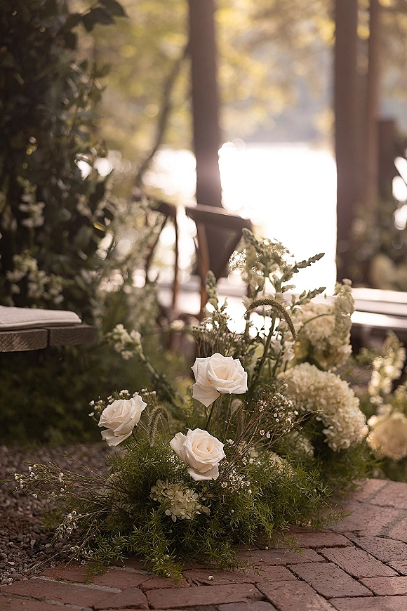 Ceremony aisle flowers with white roses and hydrangea nestled in greenery along a brick walkway in a sunlit outdoor garden setting