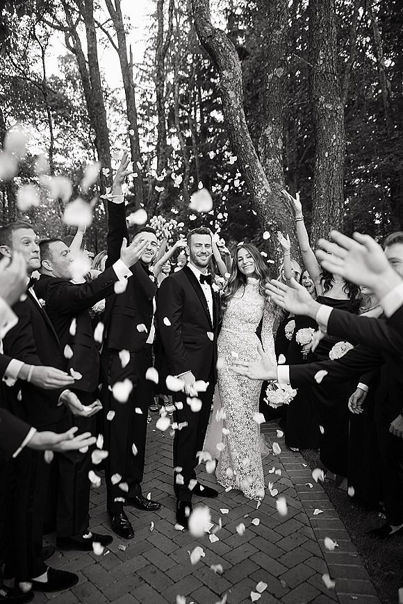 Wedding exit with rose petal toss as bride in long sleeve gown and groom in tuxedo walk brick path beneath woodland trees, guests cheering