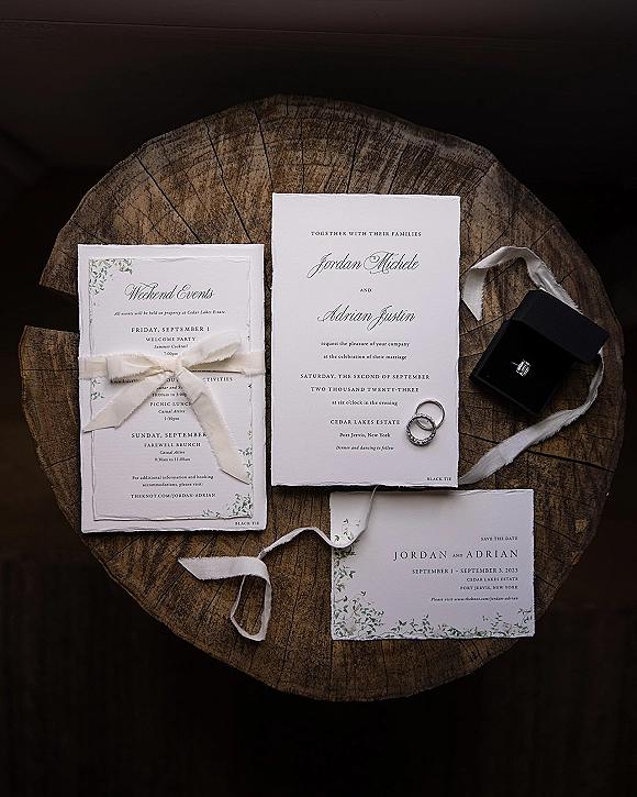 Invitation suite with deckle edge wedding invitation tied with ivory ribbon, rings and ring box beside cards on a rustic wood slice flat lay