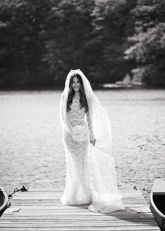 Bridal portrait of a bride holding bouquet on a dock in a lace gown with long veil, with lake, trees, and boats behind her