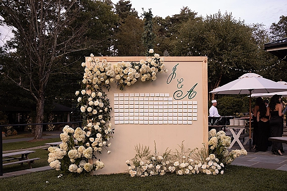 Seating chart display with wedding seating chart board and escort cards, framed by white roses and hydrangeas, greenery, and string lights on a patio lawn