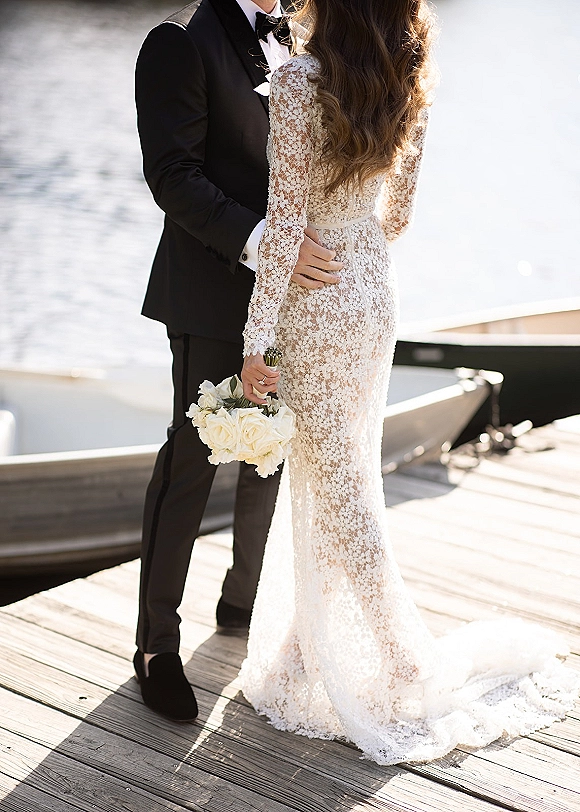 Couple portrait of bride and groom embrace on a wooden dock by the water, her lace long-sleeve gown and white rose bouquet beside a boat