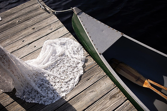 Wedding dress detail with lace wedding dress train draped on a wooden dock beside a canoe with paddles, lake water in the background