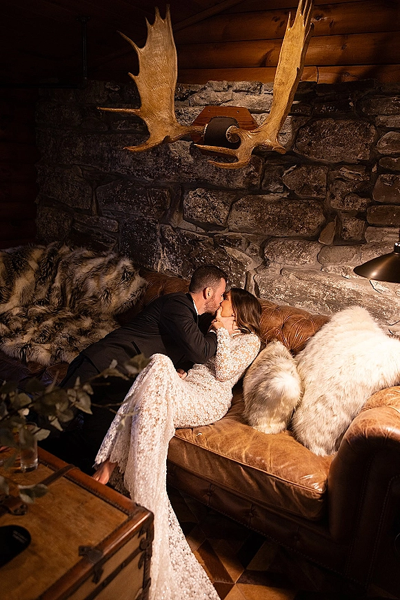 Wedding kiss portrait of bride and groom kissing on a leather sofa, bride in lace dress, in a rustic cabin with stone wall and antlers