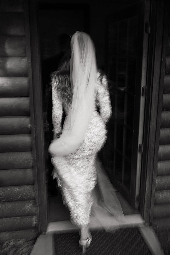 Bridal portrait of a back view bride in a lace wedding dress, veil trailing as she steps into a wooden cabin doorway in black and white