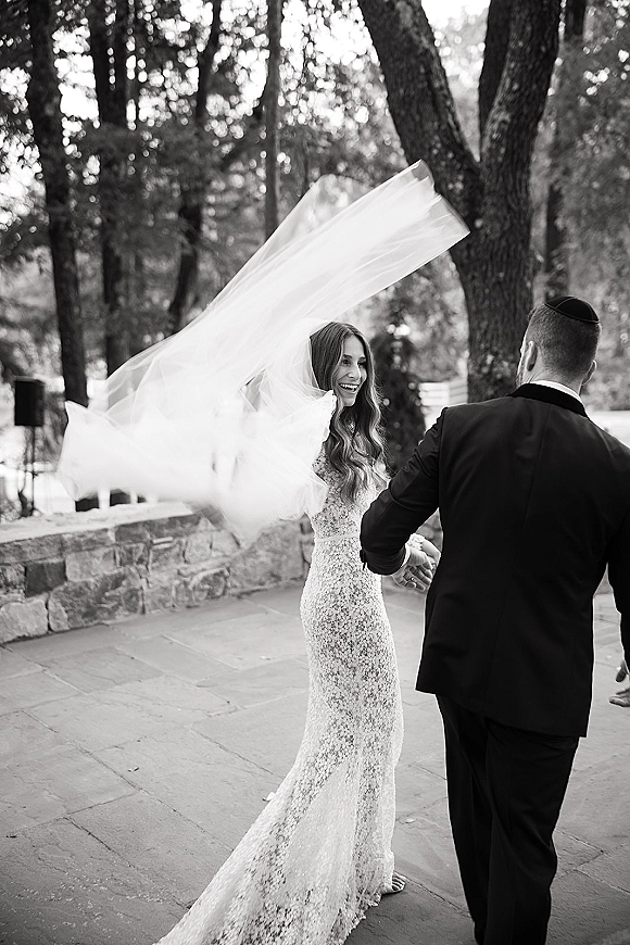 Couple portrait of bride and groom dancing, her veil blowing as he holds her hand on a stone patio with trees and wall behind