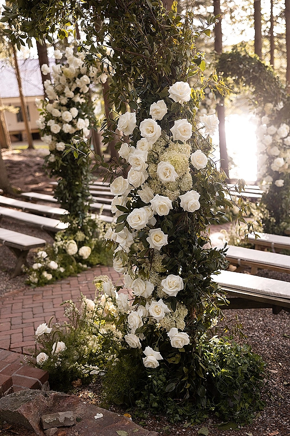 Ceremony floral arch with white roses and hydrangea, greenery garland, and floral pillars lining a brick aisle with wooden benches outdoors