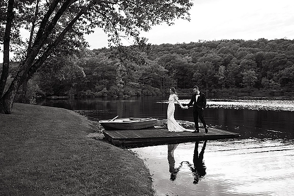 Couple portrait of bride and groom holding hands, walking on a dock by a calm lake, her lace gown train trailing beside a rowboat
