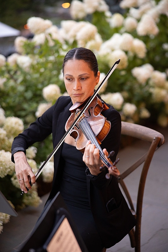 Wedding violinist in a black dress plays violin and bow beside a music stand, framed by white florals, greenery, and bistro lights outdoors