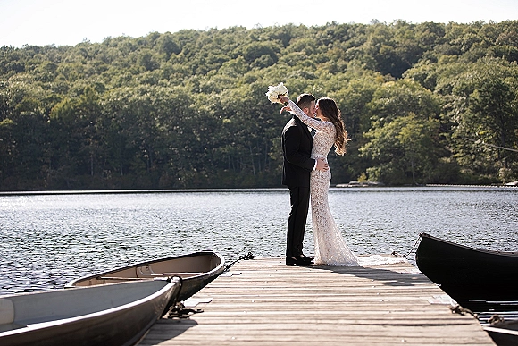 Wedding kiss portrait of bride and groom kissing on a dock, bride raising a bouquet, with a calm lake and forest shoreline behind