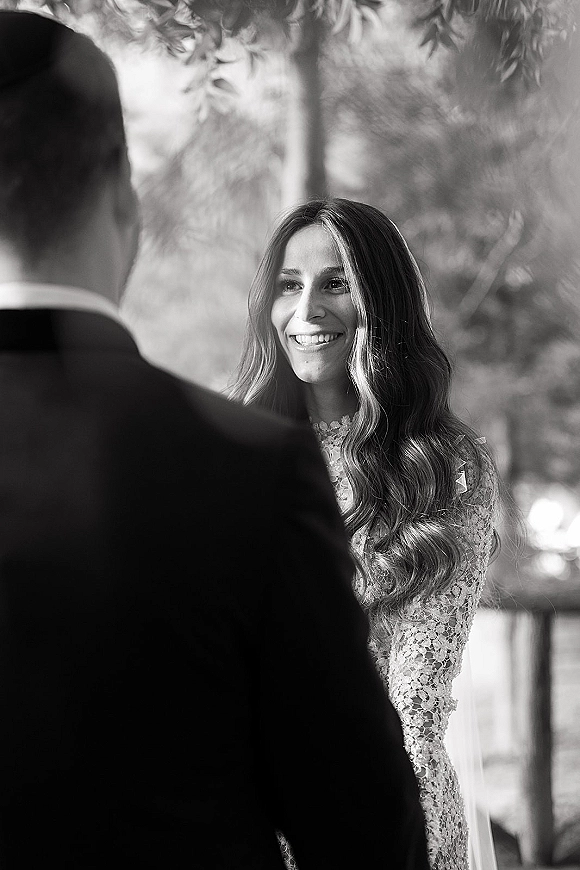 Wedding vows as bride smiles at groom, wearing a high-neck lace dress and long veil, with groom in tuxedo under leafy trees