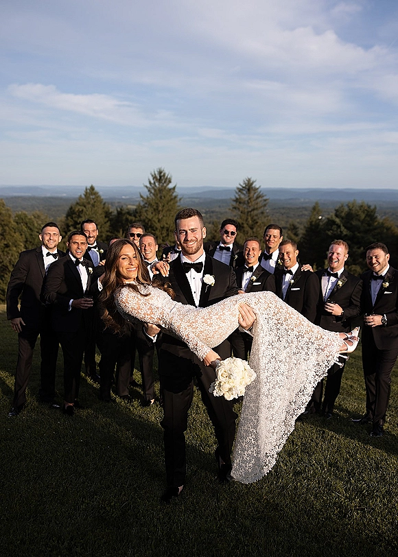 Wedding party photo of bride and groom with groomsmen, groom holding bride as the group poses on a lawn with trees and hills behind