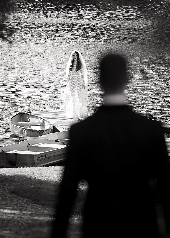 First look moment as bride in lace wedding dress and veil walks down a dock toward groom, with a rowboat and oar by the lake shore