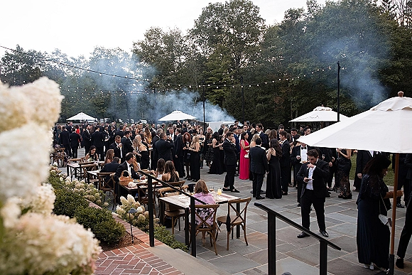 Wedding cocktail hour with black tie guests mingling under string lights, enjoying cocktails at patio tables with florals and candlelight haze