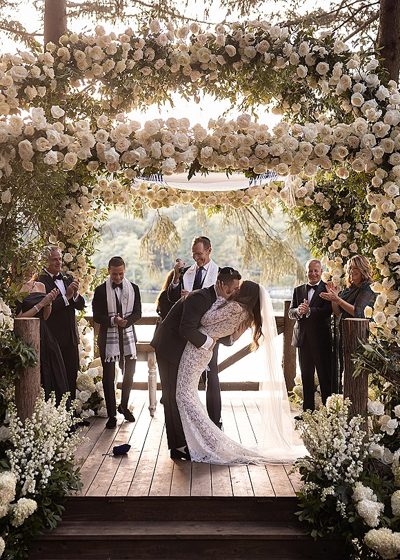Wedding kiss during ceremony kiss moment as groom dips bride in lace gown beneath white rose chuppah on a lakeside deck at sunset