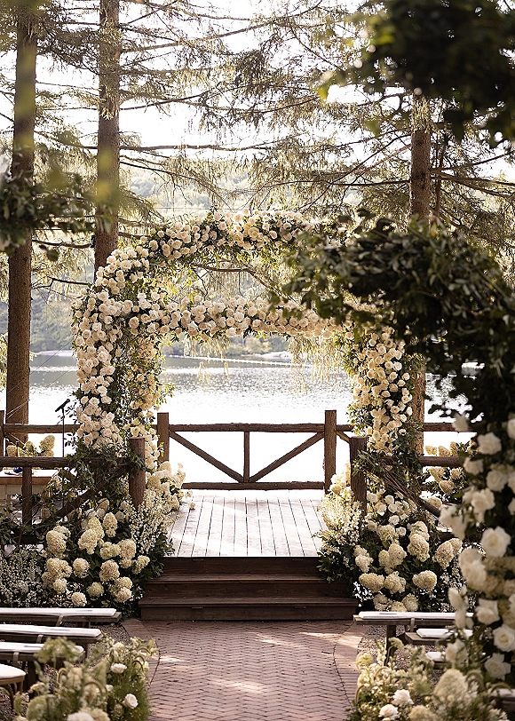 Ceremony arch draped in a floral wedding arch of white hydrangeas, roses, and greenery on a wooden deck overlooking a lake and forest hills