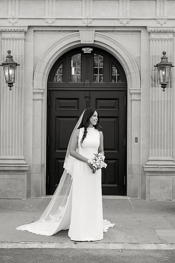 Bridal portrait in black and white of a bride holding bouquet, wearing a long veil and high neck dress by an arched doorway with columns