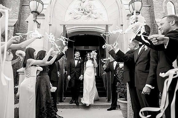 Wedding recessional with newlyweds walking out church, guests waving ribbon wands on stone steps beneath a brick arched doorway