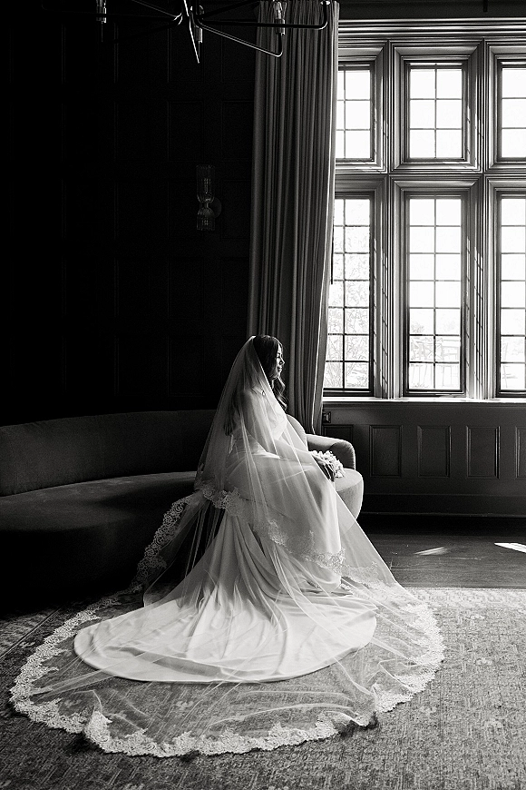 Bridal portrait in black and white of a bride seated on a curved sofa by large windows, holding a bouquet, veil and long lace train in light