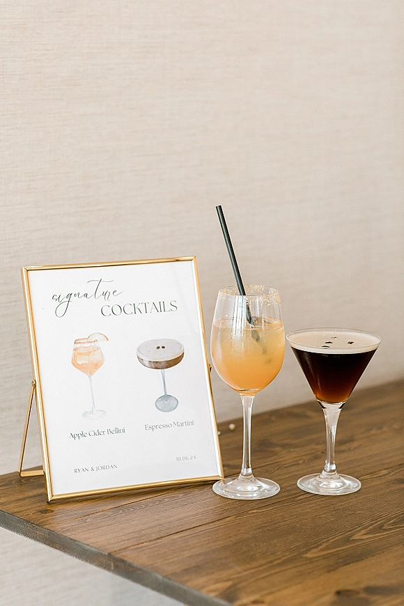 Signature cocktail sign in a gold frame with printed wedding signature cocktails menu beside glasses and black straw on wooden tabletop