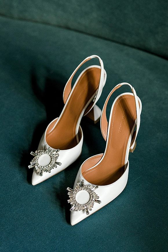 Bridal shoes, white wedding heels with jeweled shoe clips and pointed toes resting on a teal velvet sofa for a wedding day detail shot