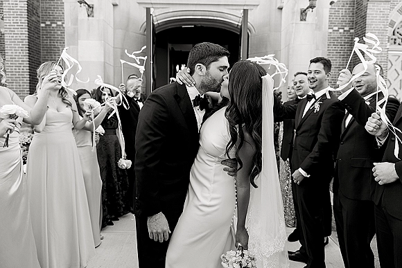 Wedding kiss portrait of newlyweds kissing at a brick church entrance, bride in strapless gown with long veil and bouquet as guests wave ribbon wands