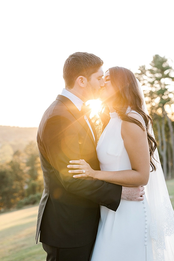 Wedding kiss portrait of bride and groom kissing at sunset, her veil blowing as golden light flares over trees and rolling hills