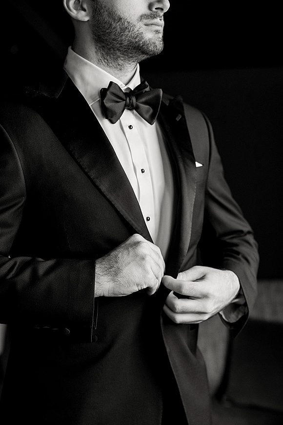 Groom portrait in a black tuxedo with bow tie, adjusting jacket buttons against a dark indoor background for a moody look