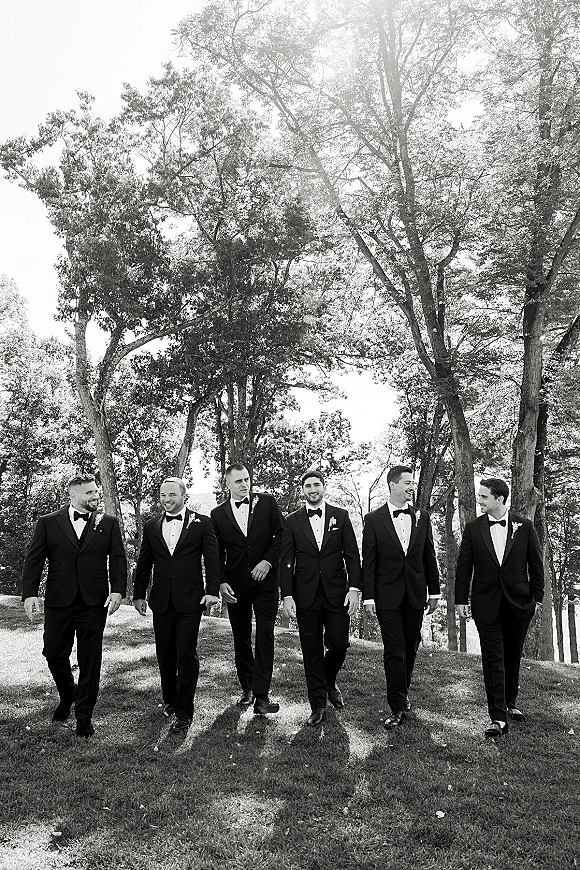 Groomsmen portrait of the wedding party in black tuxedos with bow ties, walking across a sunlit park lawn with trees behind