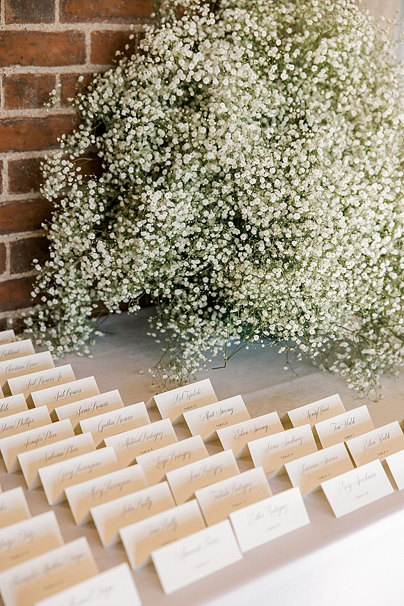 Wedding place cards in cream with calligraphy place cards and baby’s breath accents arranged on a linen tabletop against a brick wall