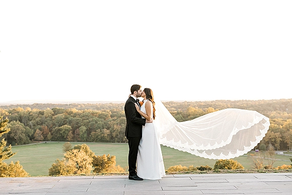 Wedding kiss portrait of bride and groom kissing as her veil blows in the wind on a stone terrace with a hilltop overlook behind them