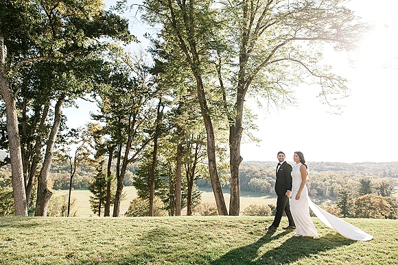 Couple portrait of bride and groom holding hands, walking through a sunlit grassy field with rolling hills, her dress train flowing