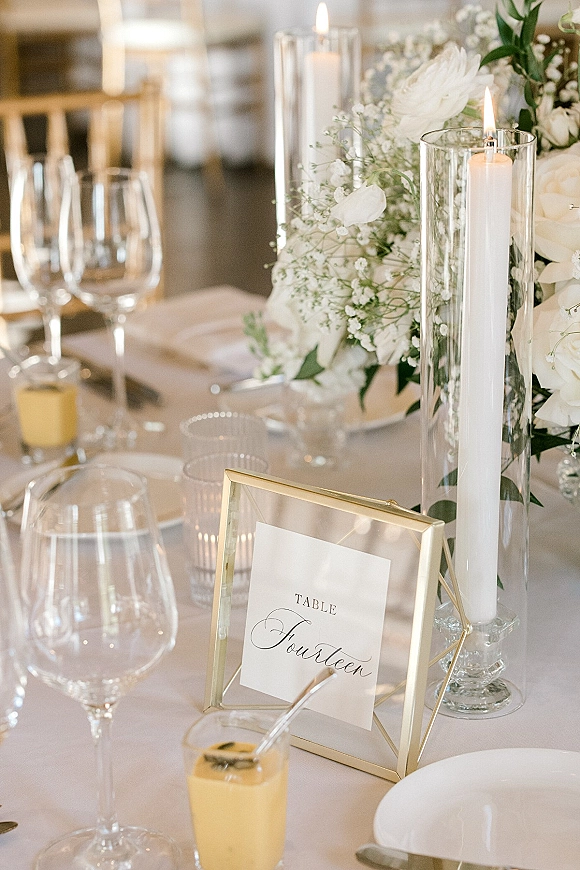 Reception tablescape with wedding table centerpiece of white florals and greenery, set with taper candles, glassware, and gold table number sign