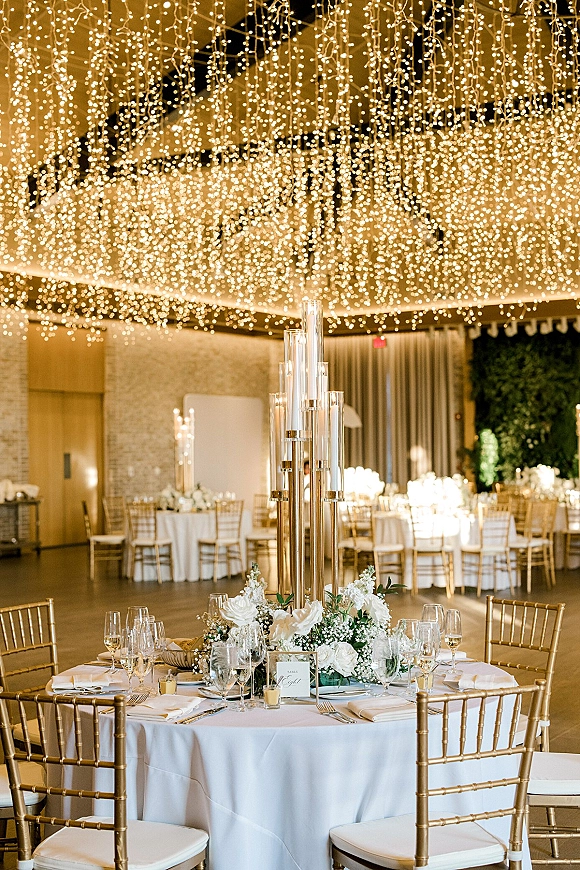 Reception tablescape with wedding string light ceiling, tall glass candles and white rose centerpiece on a round table in a draped banquet hall